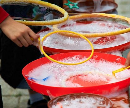 Auf dem Foto sind große, rote Schüsseln mit Seifenlauge zu sehen, in die Kinder gelbe Ringe zum Seifenblasen machen tauchen.