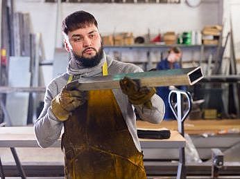 Ein Mann steht in einer Werkstatt und  hält eine Metallstange in der Hand und überprüft optisch den Werkstoff