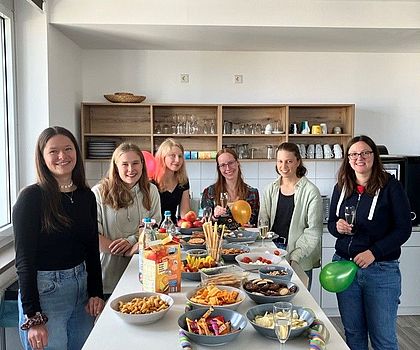 Sechs junge Frauen stehen um einen größeren Stehtisch auf welchem Snacks stehen. Sie lachnen alle in die Kamera und einigen halten einen Luftballoon in der Hand. 