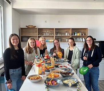 Sechs junge Frauen stehen um einen größeren Stehtisch auf welchem Snacks stehen. Sie lachnen alle in die Kamera und einigen halten einen Luftballoon in der Hand.