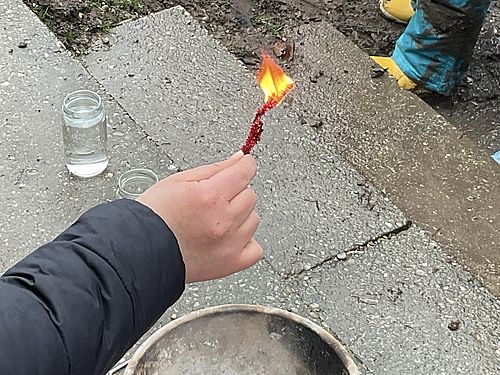 Auf diesem Bild ist eine Hand von einem Kind auf der Terrasse der Kita zu sehen, das einen brennenden Klimmstängel in der Hand hält. Auf den Terrassenfließen steht eine Schale mit einer weißen Kerze.