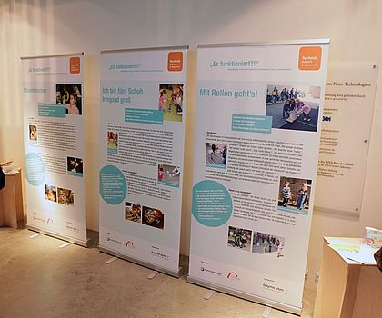 Auf dem Foto sind drei  aufgestellte Roll-up´s zu sehen, auf denen jeweils eine ausgezeichnete Projektarbeit einer Einrichtung dokumentiert ist. Die Roll-up´s stehen an einer Wand im Deutschen Museum in München und werden von geladenen Gästen zur Austellungseröffnung angeschaut.