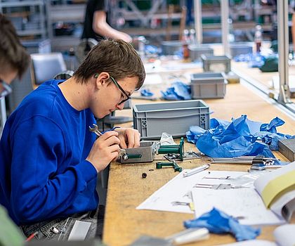 Ein Junge sitzt an einer Werkbank. Vor ihm liegt ein Roboter aus Metall. Er zieht eine Schraube mit einer Zange fest