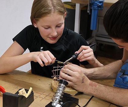 Ein Mädchen lötet einen Würfel. Ein Junge unterstützt sie dabei und hält die Drähte