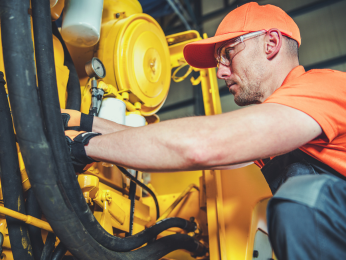 Ein Land- und Baumaschinenmechatroniker in orangefarbener Arbeitskleidung Wartung an einer gelben Baumaschine durch.
