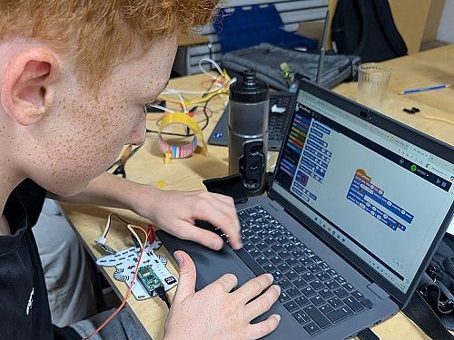 Nahaufnahme eines rothaarigen Jugendlichen von der Seite, der an einem Laptop arbeitet. Auf dem Bildschirm ist eine grafische Programmieroberfläche mit blauen Code-Blöcken zu sehen. Links neben dem Laptop liegt eine kleine, verkabelte Leiterplatine auf dem Holztisch.