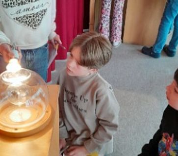 Drei Grundschulkinder knien und stehen an einer hölzernen Experimentierstation und beobachten aufmerksam eine leuchtende Glühbirne unter einer Glaskuppel. 