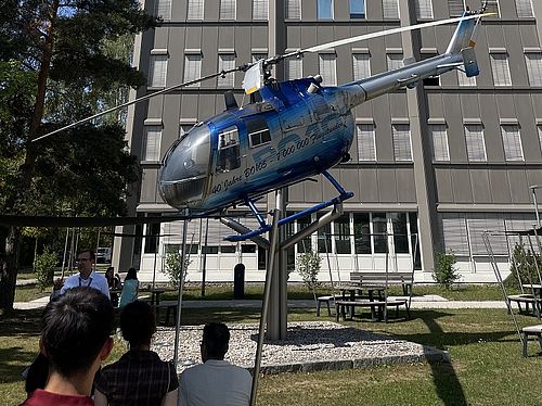 Ein blau-silberner Hubschrauber ist im Außenbereich vor einem mehrstöckigen Bürogebäude auf einer Halterung ausgestellt.