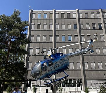 Ein blau-silberner Hubschrauber ist im Außenbereich vor einem mehrstöckigen Bürogebäude auf einer Halterung ausgestellt.