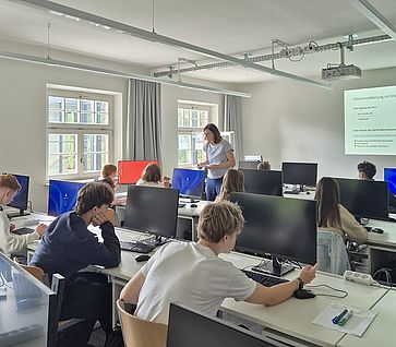 Schüler im Computerraum der TH Ingolstadt beim Planspiel Klimasimulation, eine Lehrerin steht im Hintergrund, Projektion auf Leinwand zeigt Klimamodellierung mit EN-ROADS.