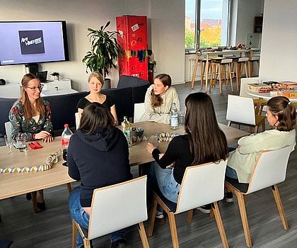 Es sitzen sechs Personen um einen Tisch. Drei sieht man von vorne, drei von hinten. Sie unterhalten sich. 