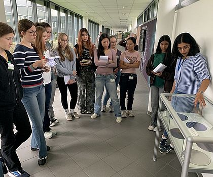 Ein Gruppe von Mädchen hört einer Frau zu. Sie stehen vor einer Vitrine