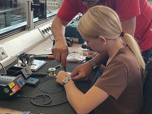 Ein Mann zeigt einem Mädchen, welches an der Werkbank sitzt, was sie beim Löten beachten muss