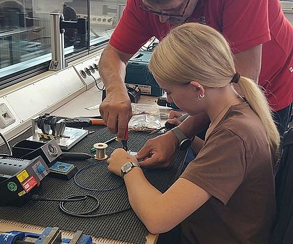 Ein Mann zeigt einem Mädchen, welches an der Werkbank sitzt, was sie beim Löten beachten muss