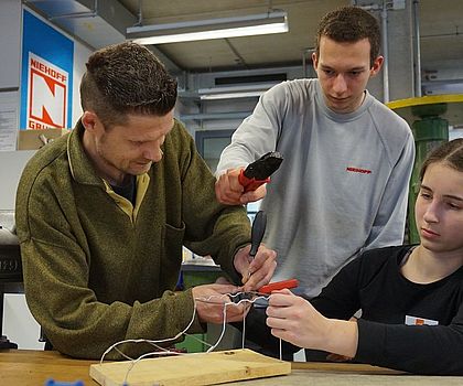 Zwei Männer helfen einem Mädchen beim Zusammenbauen der Drähte für das Werkstück.