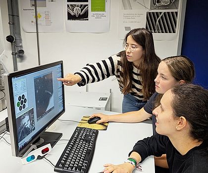 Drei Mädchen sitzen an einem Tisch. Vor Ihnen steht ein Bildschirm. Auf dem Bildschirm sieht man Zellen einer Pflanze 