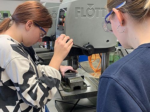 Ein Mädchen steht vor einer Standbohrmaschine und bohrt Löcher in eine Metallplatte. Ein weiteres Mädchen steht daneben.
