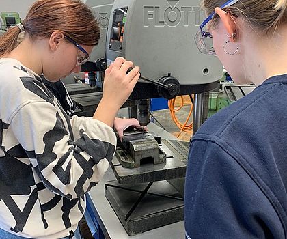 Ein Mädchen steht vor einer Standbohrmaschine und bohrt Löcher in eine Metallplatte. Ein weiteres Mädchen steht daneben.