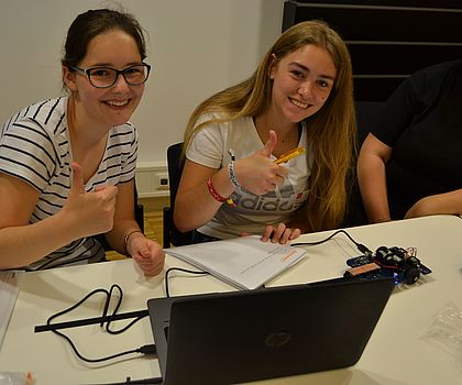 Man sieht zwei Mädchen, die den Daumen hoch zeigen und vor einem Laptop sitzen, da sie gerade coden. 