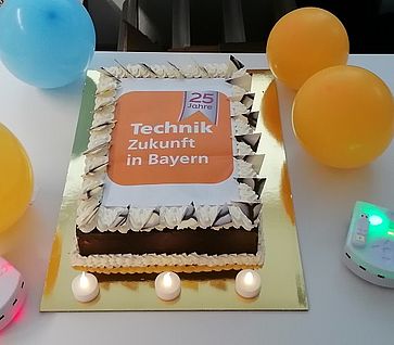 Ein Kuchen mit TeZBa-Logo. Um den Kuchen sind 4 luftballons platziert sowie zwei Thymio-Roboter die grün und rosa leuchten