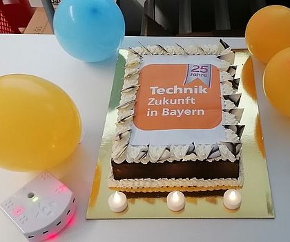 Ein Kuchen mit TeZBa-Logo. Um den Kuchen sind 4 luftballons platziert sowie zwei Thymio-Roboter die grün und rosa leuchten