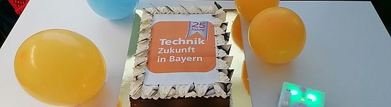 Ein Kuchen mit TeZBa-Logo. Um den Kuchen sind 4 luftballons platziert sowie zwei Thymio-Roboter die grün und rosa leuchten