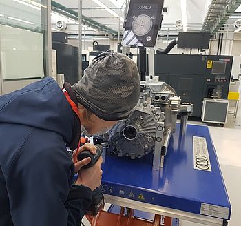 DigiCamp bei AUDI Ein junge mit Mütze und blauen Pullover steht mit einem Bohrer vor eiem Motorblock und reinigt die Zylinder