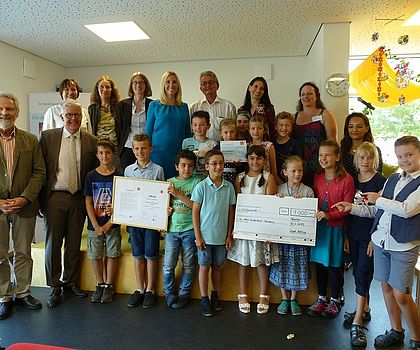 Auf dem Foto sind viele Kinder mit einem großen Check aus Pappe mit der Nennung des Preisgeldes und Urkunden zu sehen.  Die Ehrengäste sind auch mit auf dem Foto.