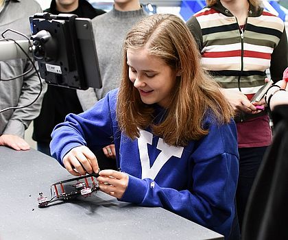 Ein Mädchen schraubt eine Drohne zusammen. Im Hintergrund sieht man weitere Mädchen, die sie beobachten.