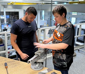 Ein Junges steht vor einem Schraubstock und feilt ein Metallstück. Neben ihn steht ein Mann und beobachtet alles