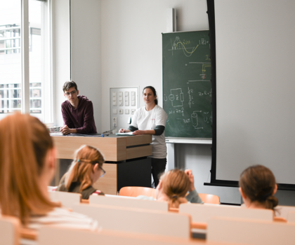 Man sieht einen Vorlesungssaal von hinten und ein paar Menschen bzw. deren Köpfe von hinten. Vorne stehen zwei Menschen hinter einem Pult.