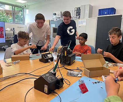 5 Jungen stehen und sitzen um einen runden Tisch und verlöten die Drähte an dsas Gehäuse ihres Werkstückes