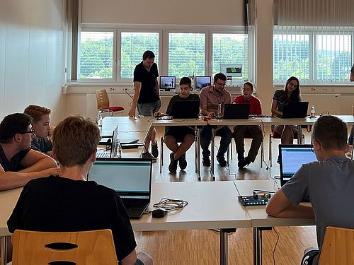 Mehrere Jugendliche sitzen an Tischen mit Laptops und programmieren Boards unter Anleitung eines Betreuers.