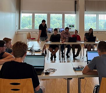 Mehrere Jugendliche sitzen an Tischen mit Laptops und programmieren Boards unter Anleitung eines Betreuers.