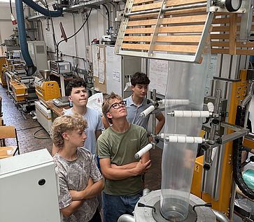 Vier Jugendliche beobachten aufmerksam einen Blasfolienextruder in einer technischen Werkstatt.