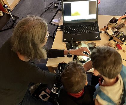 Auf dem Fotos sieht man die Workshopleiterin Ingrid Nolting und zwei Kinder, die mit einem USB-Mikroskop Steine untersuchen.