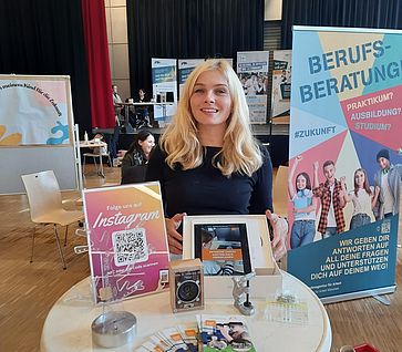 Eine Mitarbeiterin präsentiert den Stand auf der Berufsmesse. Auf dem Stehtisch vor ihr sind Flyer, Werkstücke und ein Poster mit QR Code zu sehen.
