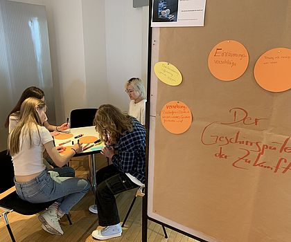 Man sieht im Vordergrund eine Pinnwand auf der steht "Geschirrspüler der Zukunft". Im Hintergrund sieht man vier Mädchen um einen runden Tisch sitzen, die etwas aufschreiben. 