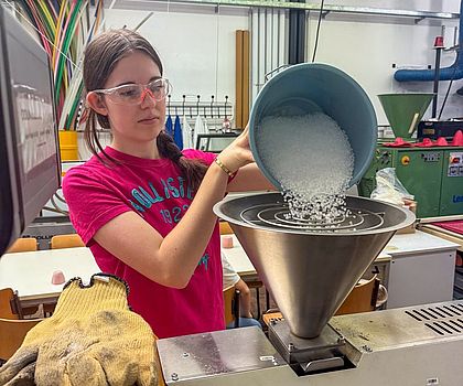 Ein Mädchen steht vor einem Trichter und füllt Plastikgranulat ein. Sie trägt eine Schuztzbrille