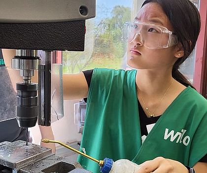 Ein Mädchen sthet vor einer Standbohrmaschine und bohrt Löcher in die Metallplatte