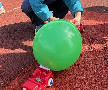 Auf dem Foto ist ein kleines, rotes Spielzeugauto zu sehen, an das ein grüner, mit Luft gefüllter Luftballon gehängt wird.