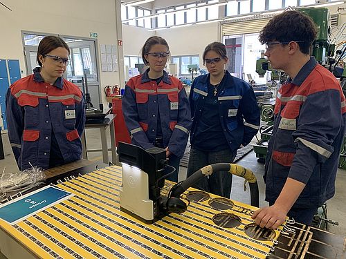 Ein männlicher Azubis erklärt einer Ruppe von Mädchen, wie die Handfräse funktioniert