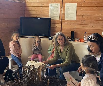 Auf dem Bild ist ein Sitzkreis um eine Baumwurzel zu sehen. Im Sitzkreis sitzen Erwachsene und Kinder. Ein Kind spricht gerade und die Aufmerksamkeit ist auf das Kind gerichtet.