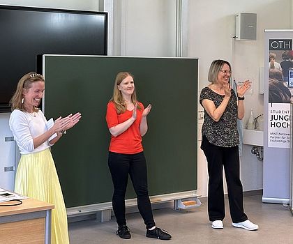 Drei Frauen stehen vor einer Tafel. Alle drei applaudieren.